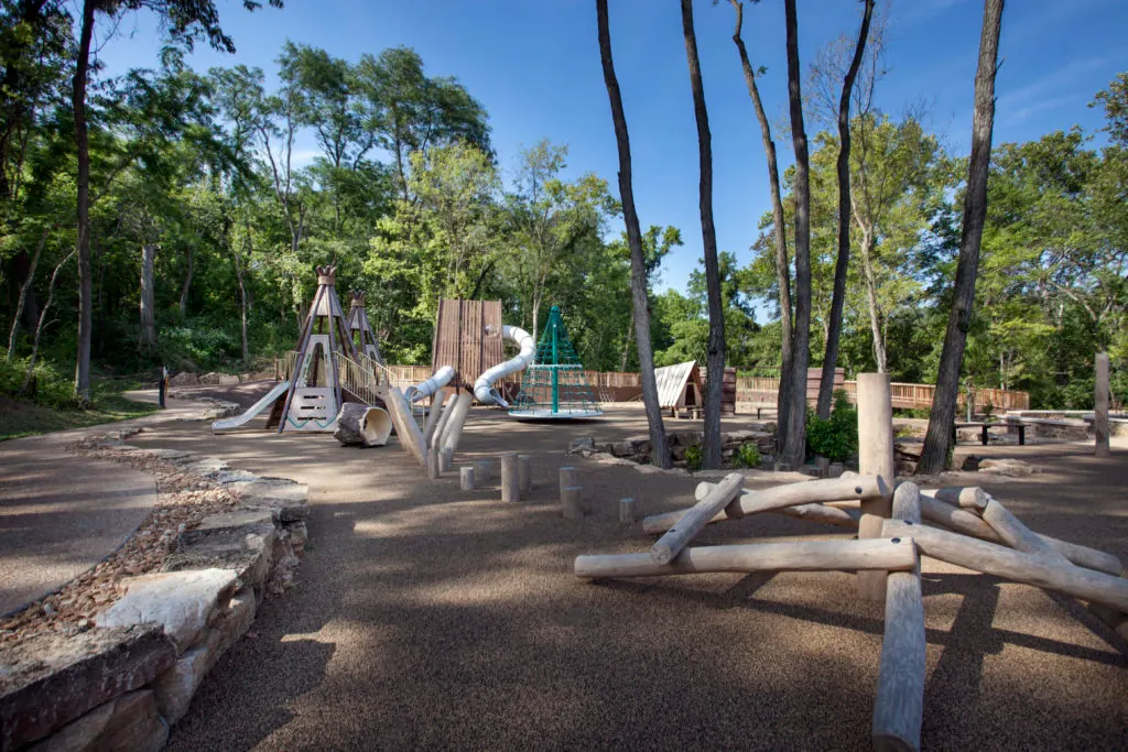 Nature-themed playground at a Wildwood, MO park with wooden structures, log climbing features, and wooded surroundings